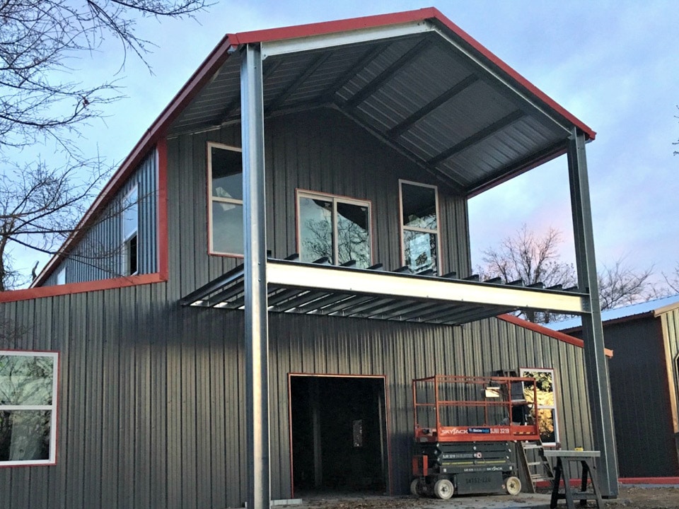 Gray steel building with red trim under construction, with a steel-framed upper porch and a scissor lift in front.